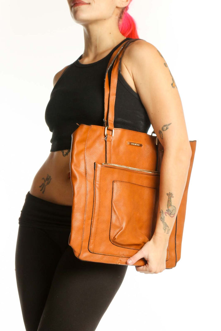 Orange leather tote bag by Steve Madden with front pocket and dual shoulder straps, held by a woman's hand