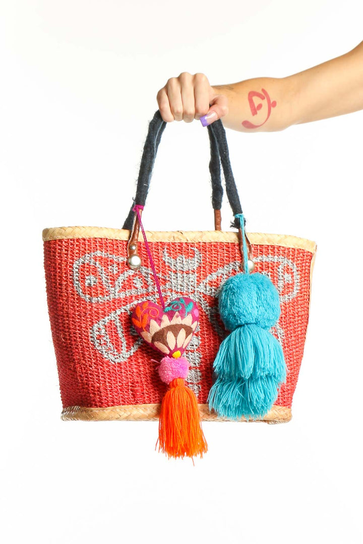 Red woven tote bag with geometric pattern, black handles, and colorful turquoise, orange, and pink tassels and pom-poms