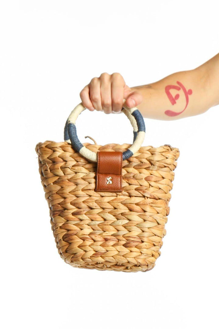Tan woven straw crossbody basket bag with cognac leather accent and navy-wrapped handles held in hand