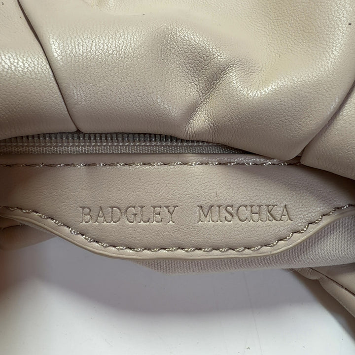 Tag view of cream Badgley Mischka shoulder bag worn on model's shoulder, displaying ruched texture and BM logo detail
