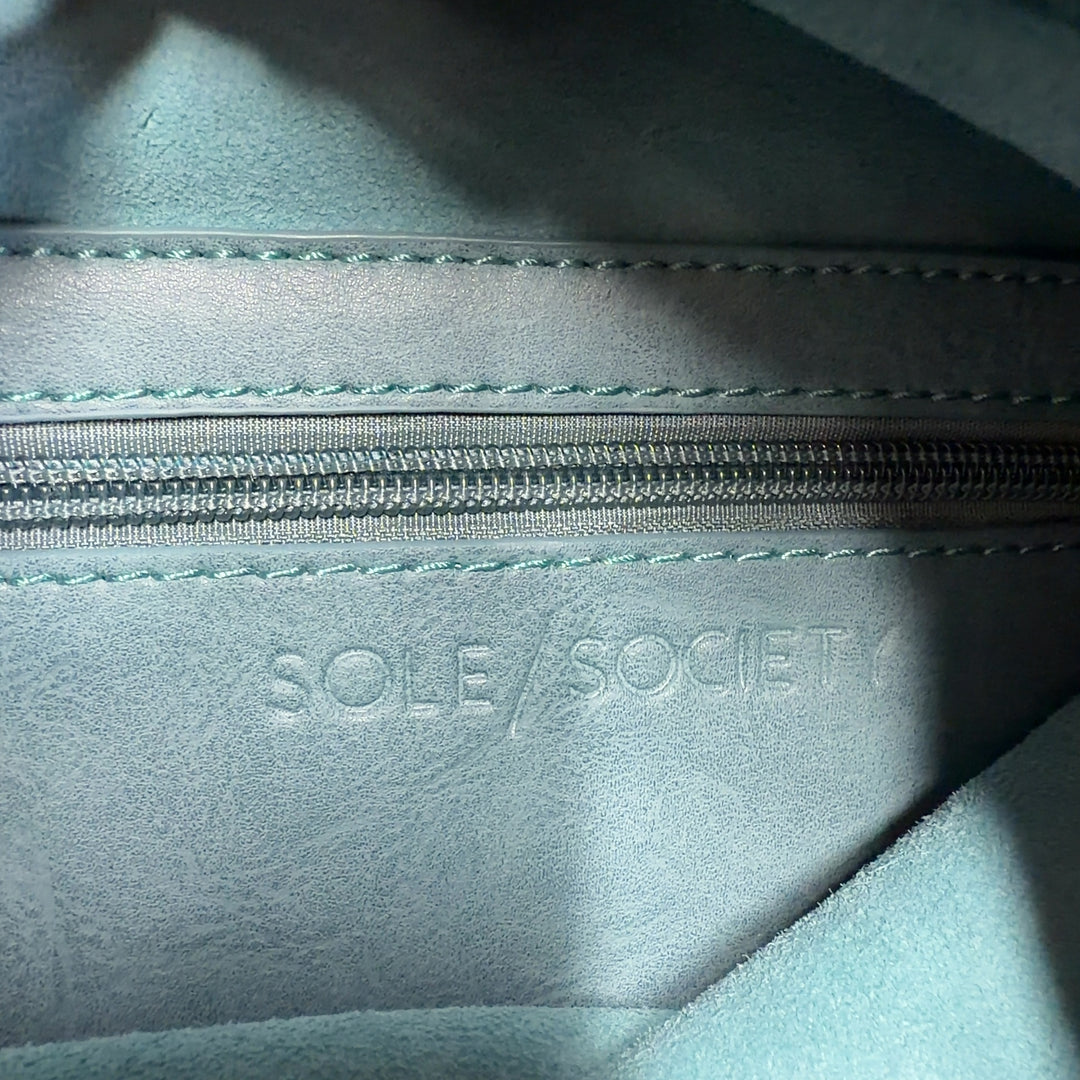 Teal suede clutch with top handle displayed from side angle, worn with black outfit