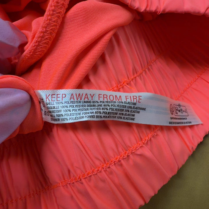 Tag view of coral pleated athletic skirt on model wearing black sports bra, displaying the flowing design and comfortable fit
